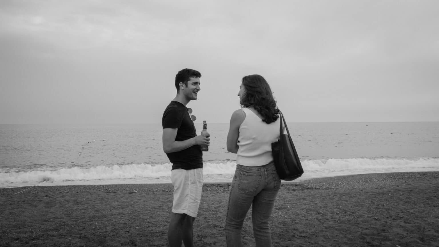 Deux personnes discutent sur une plage, l'une tenant une bouteille, face à la mer calme sous un ciel nuageux.