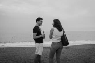 Deux personnes discutent sur une plage, l'une tenant une bouteille, face à la mer calme sous un ciel nuageux.