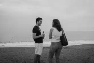 Deux personnes discutent sur une plage, l'une tenant une bouteille, face à la mer calme sous un ciel nuageux.