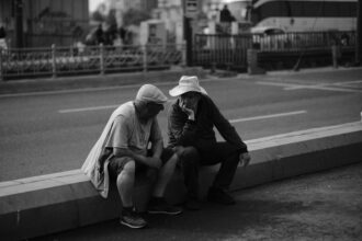 Deux hommes assis sur un trottoir discutent profondément, évoquant peut-être pourquoi les chats crachent, en pleine rue animée.