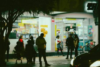 Des passants devant un magasin lumineux de nuit, discutant et utilisant leurs téléphones, créant une ambiance animée.