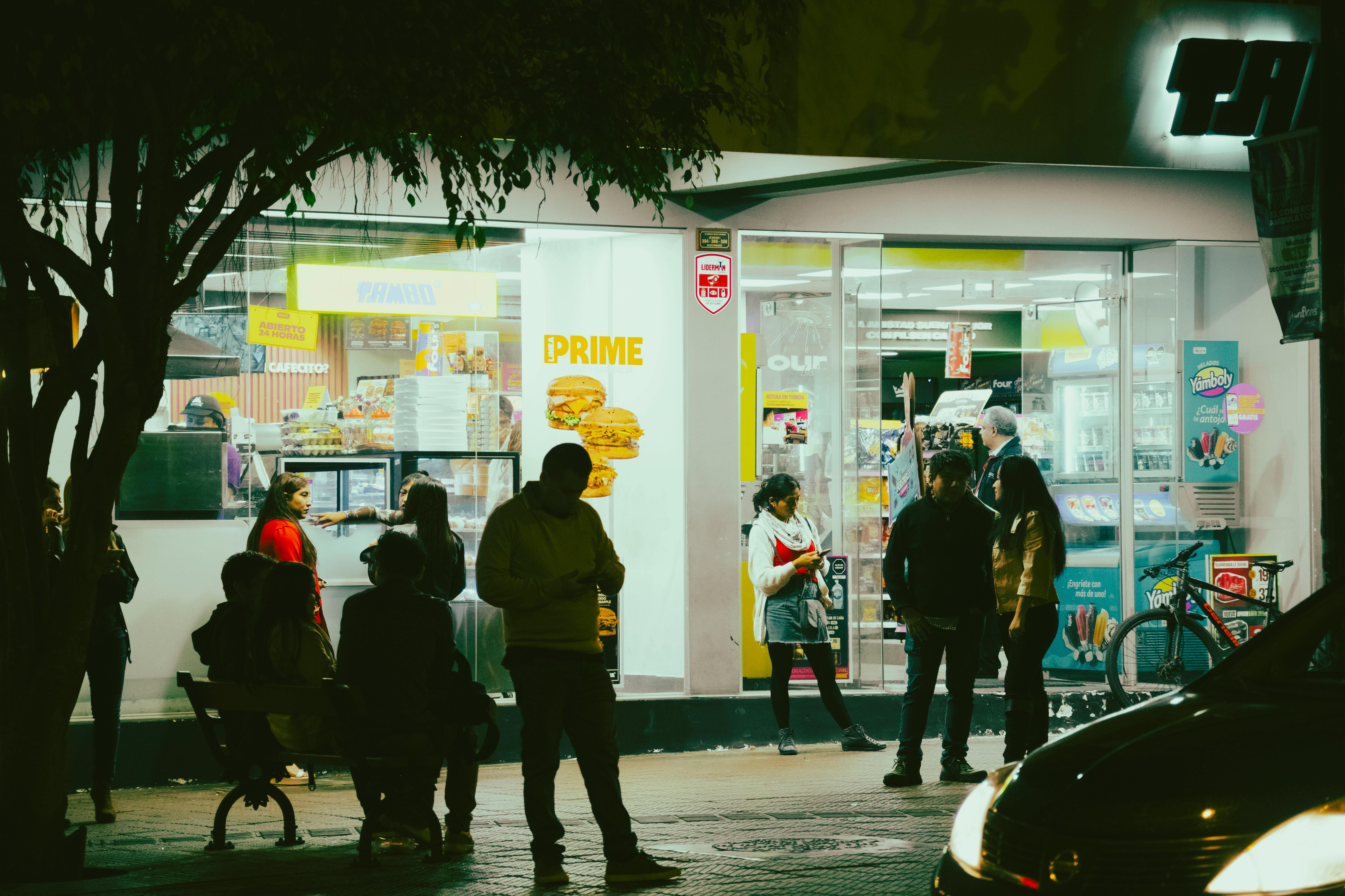 Des passants devant un magasin lumineux de nuit, discutant et utilisant leurs téléphones, créant une ambiance animée.