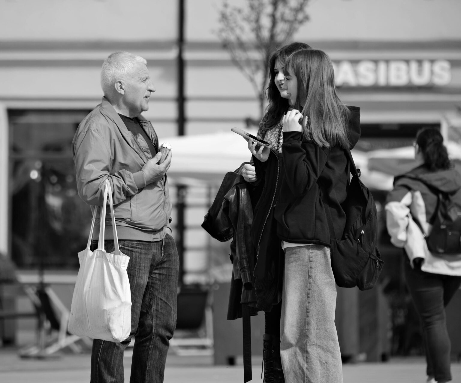 Un homme discute avec deux jeunes femmes dans la rue, souriant et tenant un sac. En arrière-plan, des passants flânent.