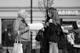 Un homme discute avec deux jeunes femmes dans la rue, souriant et tenant un sac. En arrière-plan, des passants flânent.