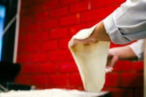 Pizzaiolo étirant une pâte à pizza devant un mur de briques rouges, évoquant pourquoi les chats patounent en pétrissant.