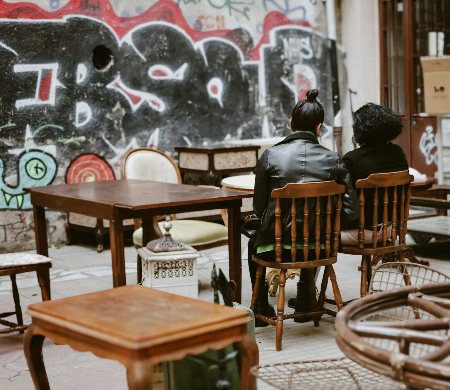 Deux personnes assises sur des chaises en bois, entourées de meubles anciens, devant un mur couvert de graffitis.