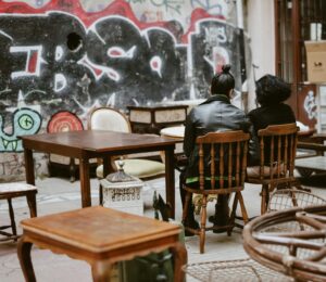 Deux personnes assises sur des chaises en bois, entourées de meubles anciens, devant un mur couvert de graffitis.