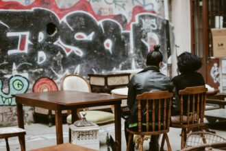 Deux personnes assises sur des chaises en bois, entourées de meubles anciens, devant un mur couvert de graffitis.