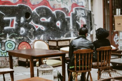 Deux personnes assises sur des chaises en bois, entourées de meubles anciens, devant un mur couvert de graffitis.