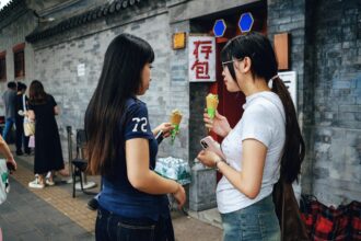 Deux femmes discutent en mangeant des glaces devant un mur gris. Une pancarte en chinois est visible à l'arrière-plan.