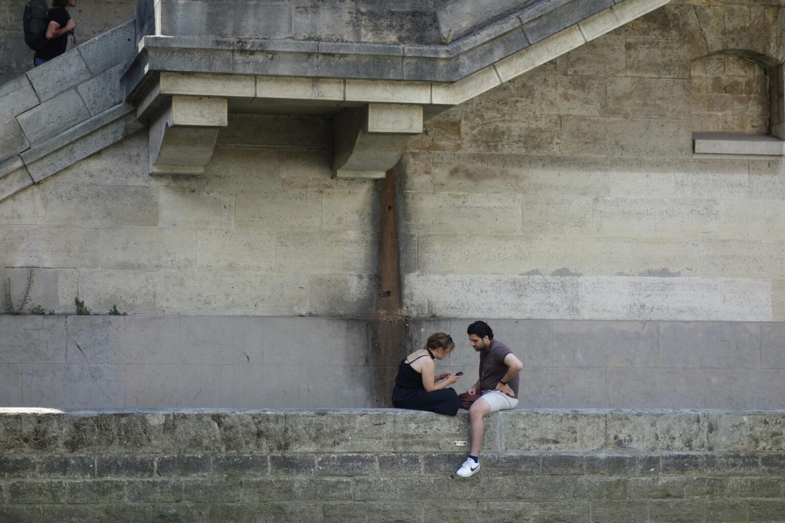 Deux personnes assises sur un muret en pierre, discutant. Un homme monte les escaliers en arrière-plan.