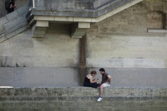 Deux personnes assises sur un muret en pierre, discutant. Un homme monte les escaliers en arrière-plan.
